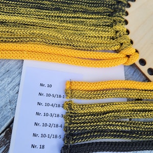May include: A color sample card featuring a gradient of braided cords ranging from solid yellow to solid black, attached to a white paper label with printed text including Nr. 10, Nr. 10-5/18-1, Nr. 10-4/18-2, Nr. 10-3/18-3, Nr. 10-2/18-4, Nr. 10-1/18-5, and Nr. 18.