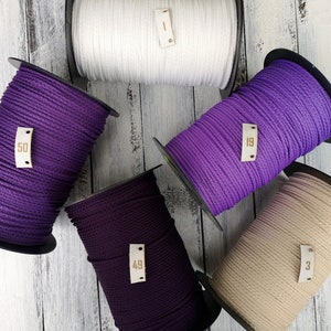 Op de afbeelding: Zes spoelen koord in verschillende kleuren, waaronder wit, paars en beige. Elke spoel heeft een klein leren label met een nummer erop. De nummers zijn 1, 19, 49, 50 en 53.