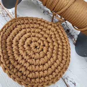 Könnte beinhalten: Ein kreisförmiges, gehäkeltes Stück aus metallischem Goldkordel mit einem dichten, spiralförmigen Stichmuster. Eine passende Spule derselben Goldkordel mit dezenten Glitzerakzenten ist direkt über dem gehäkelten Kreis positioniert.