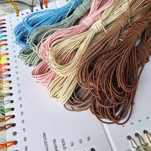 May include: A collection of colourful cords in shades of blue, pink, and brown, displayed on a white surface. The cords are bundled, and a colour chart with numbers and labels is visible. These cords are likely used for crafts or jewellery making.