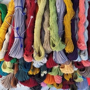Könnte beinhalten: Eine Sammlung zahlreicher Bündel aus elastischer Kordel in einer Vielzahl von Farben, darunter Rot, Gelb, Grün, Blau, Violett, Orange, Braun, Grau und Weiß. Jedes Bündel ist ordentlich aufgewickelt und mit einem kleinen Stück passender Kordel gesichert.