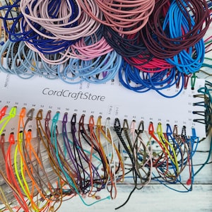 Könnte beinhalten: Eine Sammlung bunter elastischer Kordeln, die auf einer weißen Präsentationskarte mit der Aufschrift CordCraftStore angeordnet sind. Das Display zeigt zahlreiche einzelne Stränge in verschiedenen Farben, darunter Neongelb, Orange, Braun, Lila, Blau und Schwarz, mit nummerierten Schlitzen.
