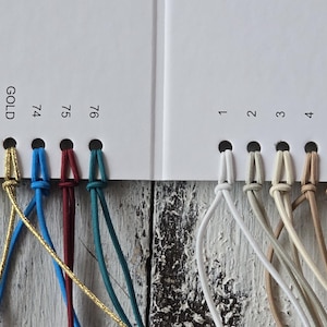 May include: A white color swatch card featuring numbered holes with attached cord samples in various colors including white, beige, blue, red, gold, silver, and black. The card includes printed labels such as REFLECTIVE, SILVER, and GOLD.