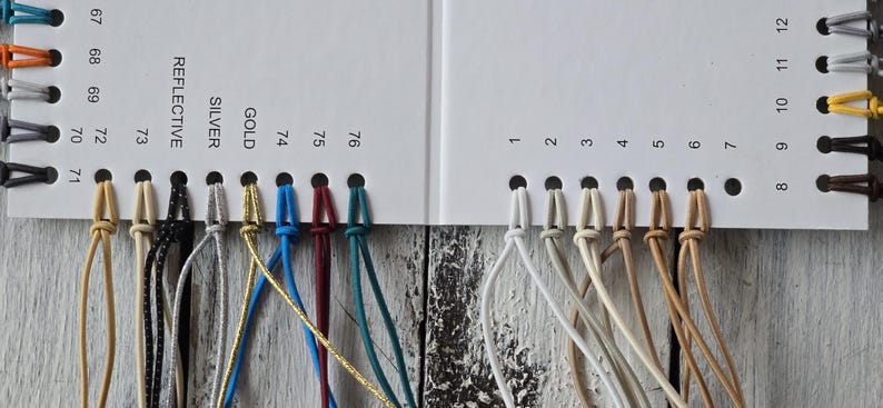 May include: A white color swatch card featuring numbered holes with attached cord samples in various colors including white, beige, blue, red, gold, silver, and black. The card includes printed labels such as REFLECTIVE, SILVER, and GOLD.