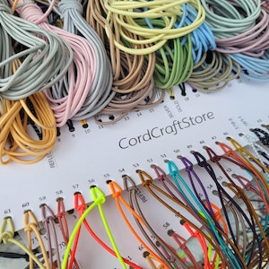 Könnte beinhalten: Eine Farbmusterkarte von CordCraftStore mit zahlreichen Bündeln dünner, mehrfarbiger elastischer Kordeln. Die weiße Karte enthält aufgedruckte numerische Etiketten und Farbnamen, wobei verschiedene Kordeln durch Löcher gefädelt sind, um ein breites Farbspektrum zu zeigen.