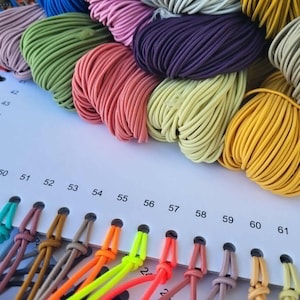 May include: A selection of colorful elastic cords in various shades, including pink, green, yellow, and purple. The cords are arranged in neat bundles, with a color chart featuring numbered samples below. The image showcases a variety of crafting supplies.