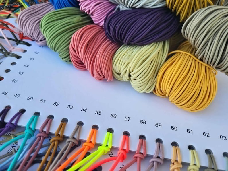 May include: A color chart featuring numerous bundles of elastic cord in various shades including yellow, green, pink, purple, and grey. The cords are attached to a white card with numbered holes ranging from 42 to 63, displaying a wide spectrum of colors.