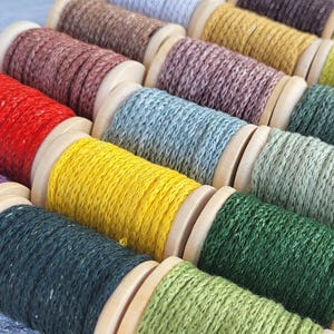Könnte beinhalten: Eine Sammlung von Holzspulen, die mit verschiedenfarbigem, strukturiertem Garn umwickelt sind, darunter Rot, Gelb, Grün, Blau, Violett, Braun und Grau. Die Spulen sind in Reihen angeordnet und zeigen eine Palette aus lebendigen und gedeckten Tönen.