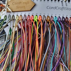 May include: A color sample card from CordCraftStore featuring a wide array of thin, flexible cords in various solid colors including neon orange, yellow, green, purple, blue, and brown, with numbered slots and a wooden brand tag attached.