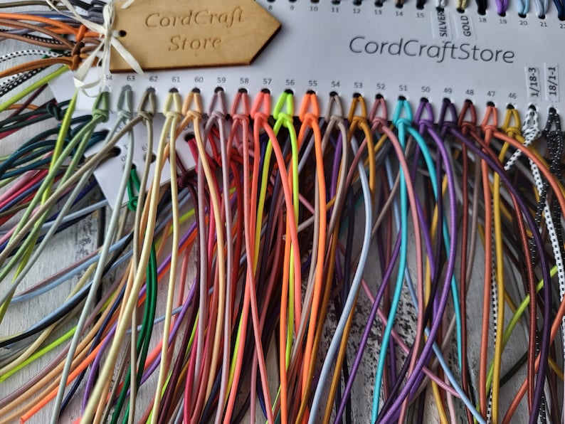 May include: A color sample card from CordCraftStore featuring a wide array of thin, flexible cords in various solid colors including neon orange, yellow, green, purple, blue, and brown, with numbered slots and a wooden brand tag attached.