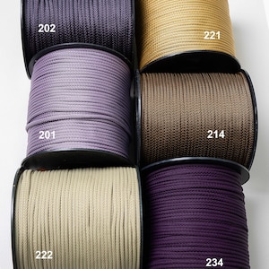 Op de afbeelding: Zes spoelen met gevlochten koord in verschillende kleuren, elk gelabeld met een nummer: 202 in donkerpaars, 201 in lichtpaars, 222 in beige, 221 in goud, 214 in bruin en 234 in dieppaars. De spoelen zijn in twee kolommen gerangschikt.