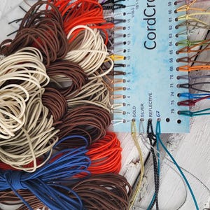 May include: A color sample card labeled CordCraftStore with numbered holes from 1 to 76, featuring various elastic cords in shades of red, brown, cream, blue, gold, silver, and reflective black, organized by color and attached to the card.