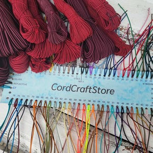 Könnte beinhalten: Eine Farbkarte mit der Aufschrift CordCraftStore, die nummerierte Löcher mit befestigten dünnen Schnüren in einer Vielzahl von Farben wie Rot, Blau, Grün, Gelb und Orange zeigt, mit großen Bündeln aus roter und burgunderroter Schnur darüber.