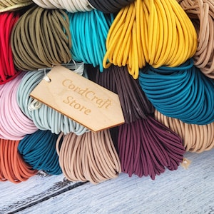Könnte beinhalten: Eine Sammlung elastischer Kordeln in verschiedenen Farben wie Rot, Gelb, Blau, Rosa, Braun und Beige, die in Bündeln angeordnet sind. Ein helles Holzschild mit der eingravierten Aufschrift CordCraft Store liegt auf den bunten Kordelbündeln.