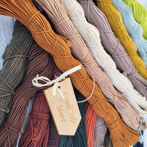 Könnte beinhalten: Eine Sammlung mehrfarbiger elastischer Kordeln, die in verschiedenen Farbtönen wie Orange, Braun, Beige, Gelb, Blau und Grün gebündelt sind. Ein helles Holzschild mit dem Text CordCraft Store ist mit einer weißen Schnur an den Bündeln befestigt.