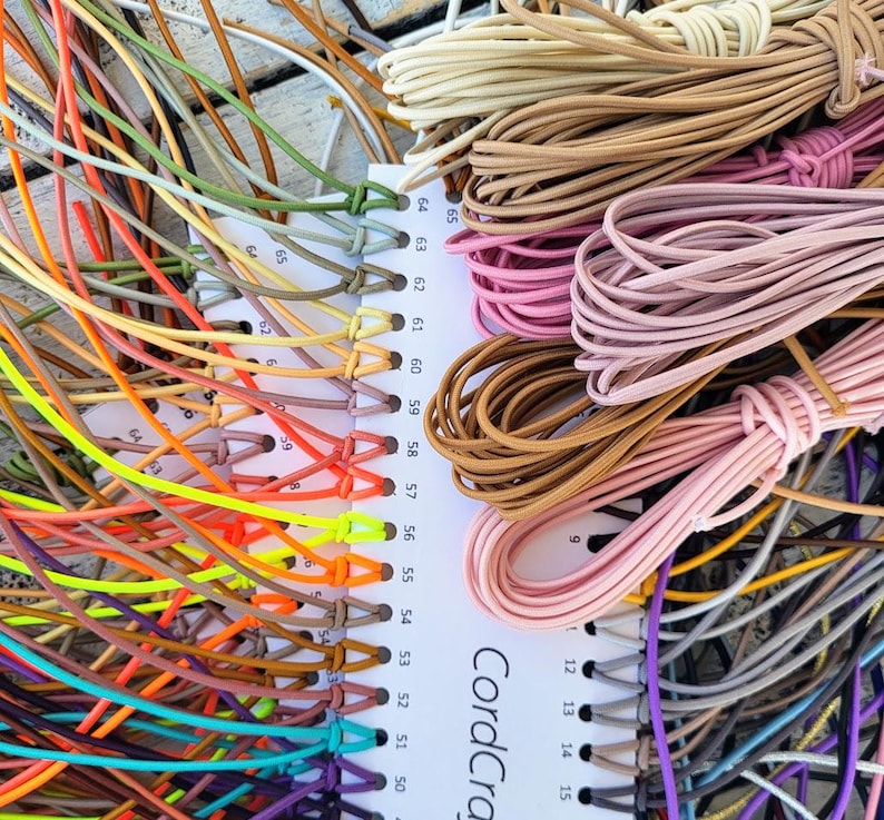 May include: A white color chart labeled CordCraft with numbered holes holding various lengths of elastic cord in a wide spectrum of colors including neon yellow, orange, pink, brown, tan, blue, purple, green, and grey, arranged in bundles.