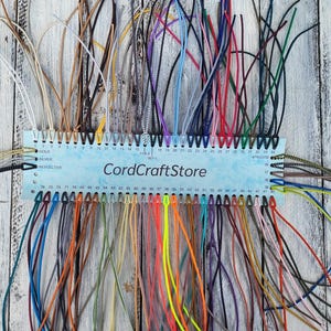 May include: A rectangular light blue display card labeled CordCraftStore featuring numerous numbered holes along the top and bottom edges. Each hole holds a thin, colorful cord in various shades including neon, metallic, and neutral tones.