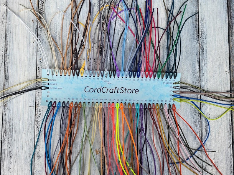 May include: A rectangular light blue display card labeled CordCraftStore featuring numerous numbered holes along the top and bottom edges. Each hole holds a thin, colorful cord in various shades including neon, metallic, and neutral tones.