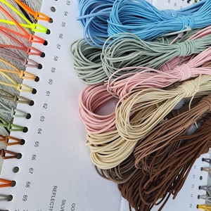 Könnte beinhalten: Eine Auswahl an farbigen Kordeln und Schnurmustern wird auf einer weißen Karte mit nummerierten Farbfeldern gezeigt. Die Kordeln sind in Blau-, Grün-, Rosa-, Beige- und Brauntönen gehalten. Die Karte enthält auch reflektierende, silberne und goldene Optionen.