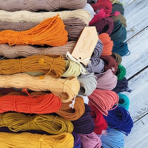 May include: A collection of numerous coiled elastic cords in a wide variety of solid colors including orange, yellow, red, purple, blue, and beige. A light-colored wooden tag with the text CordCraft Store is tied to the center of the cords.