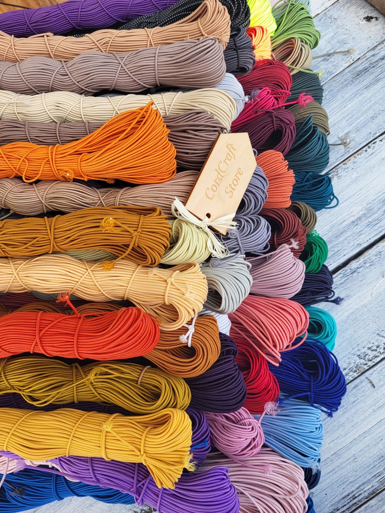 May include: A collection of numerous coiled elastic cords in a wide variety of solid colors including orange, yellow, red, purple, blue, and beige. A light-colored wooden tag with the text CordCraft Store is tied to the center of the cords.