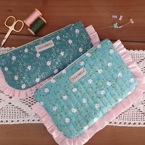 Pode incluir: Duas necessaires com estampa floral e babados rosa. As necessaires são verde-azuis com um padrão floral branco e uma etiqueta que diz "CHICA DANISCA". Materiais de costura, incluindo tesouras, carretéis de linha e alfinetes, são dispostos ao redor.