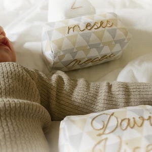 May include: Baby milestone blocks with geometric patterns in yellow, gray, and white. The blocks are embroidered with gold thread, displaying the number "2", the word "meses", and a name. The blocks are designed for baby milestones.