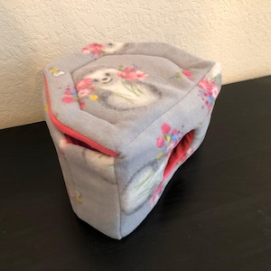 May include: A soft, gray, fleece-like pet hideout with a hexagonal shape. The fabric features a repeating pattern of sloths surrounded by pink and blue flowers. The interior of the structure is lined with a solid, vibrant coral-pink fabric.