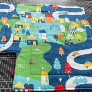 May include: A colorful play mat featuring a whimsical town scene with houses, trees, roads, and cars. The mat has a blue background with green and white accents, and a red border. The word "ATTICA" is visible on the mat.