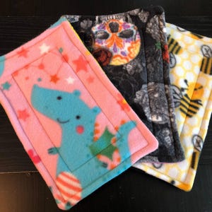 May include: Three rectangular fleece pads featuring distinct patterns. One is pink with a blue dinosaur and stars, one is black with colorful sugar skulls and roses, and one is white with a yellow honeycomb pattern and black and yellow bees.