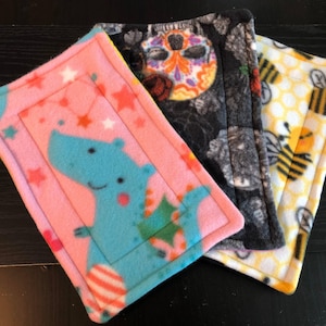 May include: Three fleece pads with different patterns. One is pink with a blue dinosaur and stars, another is black with a sugar skull design, and the third is yellow with bees. Each pad has a stitched border.