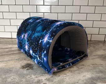 May include: A small pet tunnel bed with a galaxy print. The tunnel is primarily dark blue with white and purple starburst patterns. The interior and edges are a soft gray color. The bed is designed for small pets.