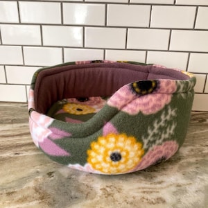 May include: A small pet bed with a floral pattern. The bed features a dark green exterior with pink and yellow flower designs. The interior is a solid mauve color, and the bed is made of a soft, plush material.