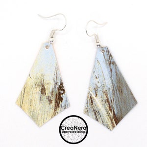 Peut inclure: Une paire de boucles d'oreilles pendantes argentées. Chaque boucle d'oreille présente un pendentif en forme de losange avec un motif texturé et vieilli dans les tons de bleu, marron et jaune. Fermeture à crochet. Le logo "CreaNera Upcycled bling" est visible.