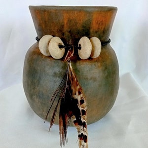 May include: A dark brown ceramic vase with a beaded trim and a single brown feather hanging from it.