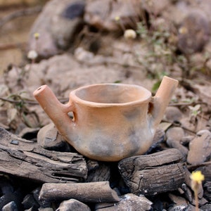 Puede incluir: Una pequeña olla de barro marrón con dos picos se asienta sobre un lecho de carbón vegetal negro. La olla tiene una superficie rugosa y texturizada y parece estar hecha a mano.