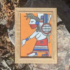 May include: A framed painting featuring a stylized figure with red, white, and blue accents against an orange background. The figure is depicted in profile, wearing a headdress and holding a circular object. The frame is made of light-colored wood.