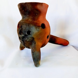 May include: A terracotta pot with a brown, orange, and black glaze. The pot has a round body with holes and three legs. It is a traditional style pot.