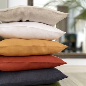 May include: A stack of five square throw pillows in various solid colors, including beige, white, mustard yellow, terracotta orange, and dark charcoal gray. Each pillow features a visible fabric texture and a simple stitched seam along the edge.