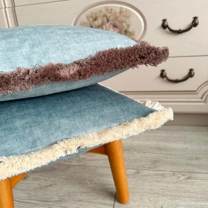 May include: Two square, light blue decorative cushions with fringe trim. One cushion has brown fringe, the other has cream-coloured fringe. The cushions are stacked on a small wooden stool with light brown legs.