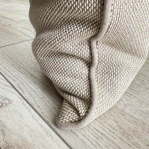 May include: Close-up of a beige throw pillow with a textured fabric and a white seam around the edge.
