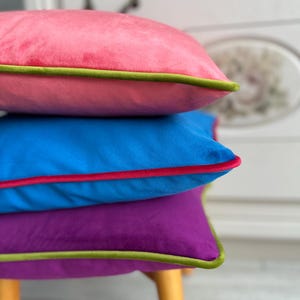 Peut inclure: Trois coussins carrés en velours rose, bleu et violet empilés. Le coussin rose a une bordure verte, le bleu une bordure rouge et le violet une bordure verte. Les coussins sont empilés sur un tabouret en bois clair.