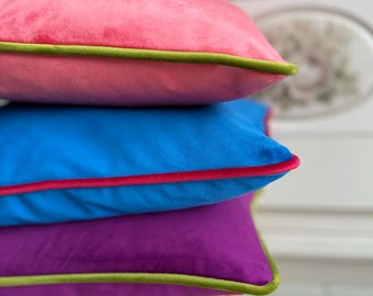 May include: A stack of three square velvet throw pillows in vibrant colors. The top pillow is pink with a green trim, the middle is blue with a red trim, and the bottom is purple with a green trim. The pillows are stacked on a light wooden stool.