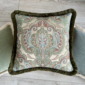 Puede incluir: Una almohada cuadrada con un patrón de paisley verde, marrón y azul. La almohada tiene un borde de flecos verde.