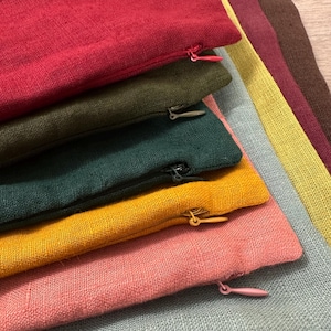 May include: A stack of rectangular fabric cushion covers featuring a visible woven texture. The covers are layered in colors including red, olive green, dark teal, mustard yellow, and pink, each with a matching zipper closure on the side.