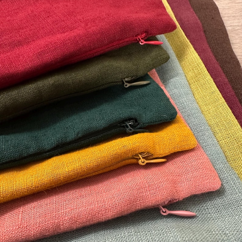 May include: A stack of rectangular fabric cushion covers featuring a visible woven texture. The covers are layered in colors including red, olive green, dark teal, mustard yellow, and pink, each with a matching zipper closure on the side.