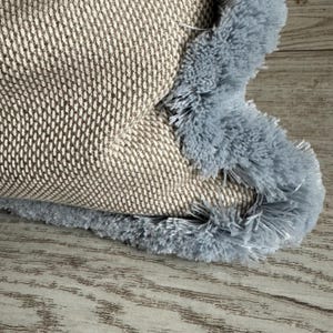 Può includere: Una federa quadrata per cuscino caratterizzata da un tessuto beige testurizzato a trama fitta. I bordi del cuscino sono rifiniti con una spessa e morbida frangia color azzurro che aggiunge un bordo decorativo al materiale neutro.