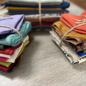 Puede incluir: Montones de cuadrados de tela coloridos atados con hilo. Las telas incluyen tonos de rojo, verde, azul y amarillo. Los paquetes están dispuestos sobre una superficie gris claro.