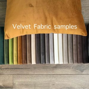 Könnte beinhalten: Eine Auswahl an Samtstoffmustern in verschiedenen Farben, darunter Grün, Gelb, Braun, Grau und Weiß. Der Text "Velvet Fabric samples" ist auf einem senffarbenen Samtkissen zu sehen. Die Muster sind in einer Reihe auf einer Holzoberfläche angeordnet.