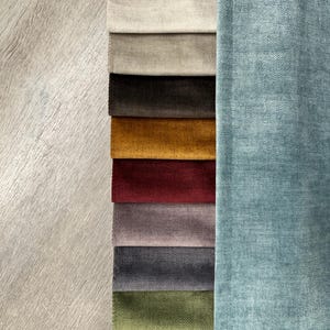 Puede incluir: Una exhibición de muestras de tela con una variedad de colores y texturas. Las muestras incluyen tonos de gris, beige, marrón, dorado, burdeos, morado y verde. Un trozo más grande de tela azul claro está a la derecha.